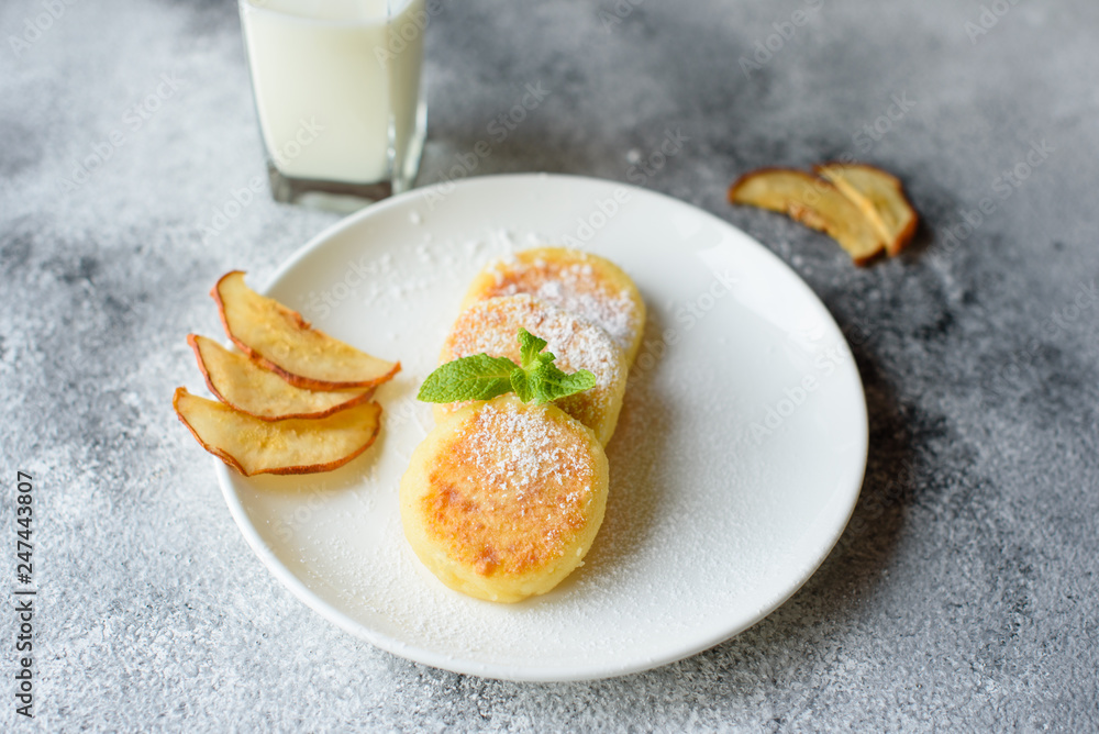 Tasty fresh cottage cheese pancakes on a white plate with a glass of milk on a concrete background. Healthy and diet breakfast