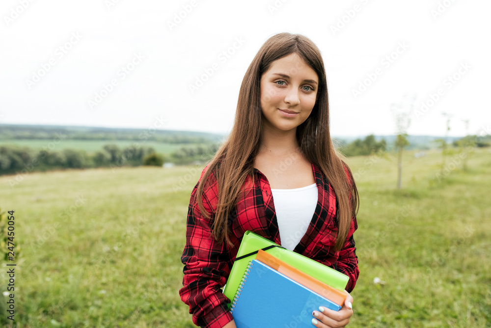 Fototapeta premium A girl teenager 10-13 years old is standing in a park in summer against background of green meadows and fields. In hands of holding notebooks and document folders in city park.