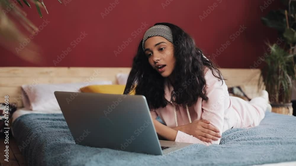 Happy african american girl lies on her belly on the bed by the window happily chatting with her friends on a video call on her laptop, slow motion Happy african american girl lies on her belly on the bed by the window happily chatting with her friends on a video call on her laptop, slow motion