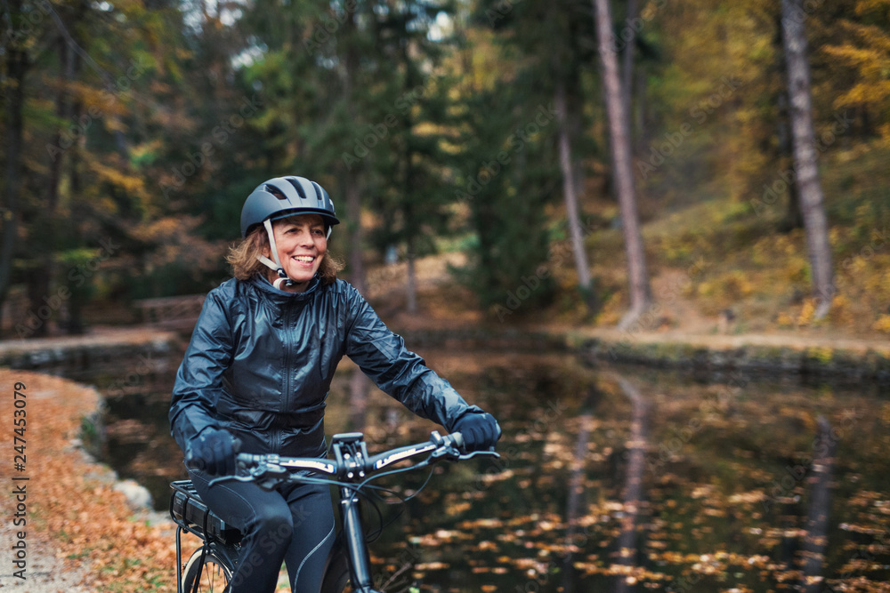 A senior woman with electrobike cycling outdoors on a road in park in autumn. A senior woman with electrobike cycling outdoors on a road in park in autumn.