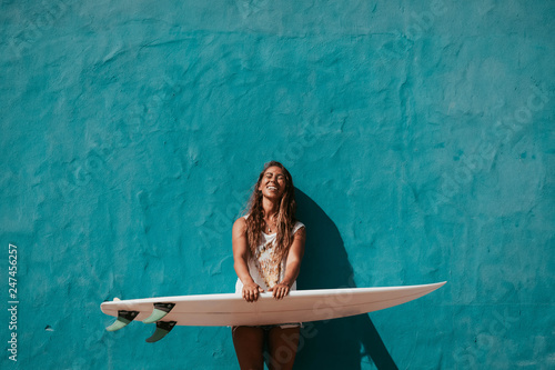 happy surfer girl with surfboard in front of blue wall