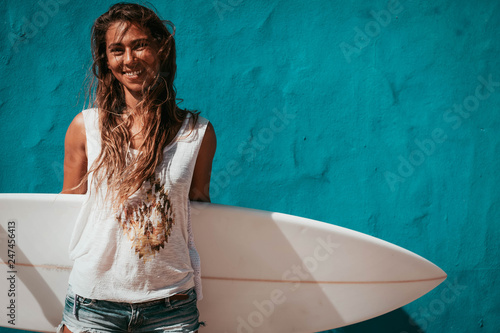 happy surfer girl with surfboard in front of blue wall