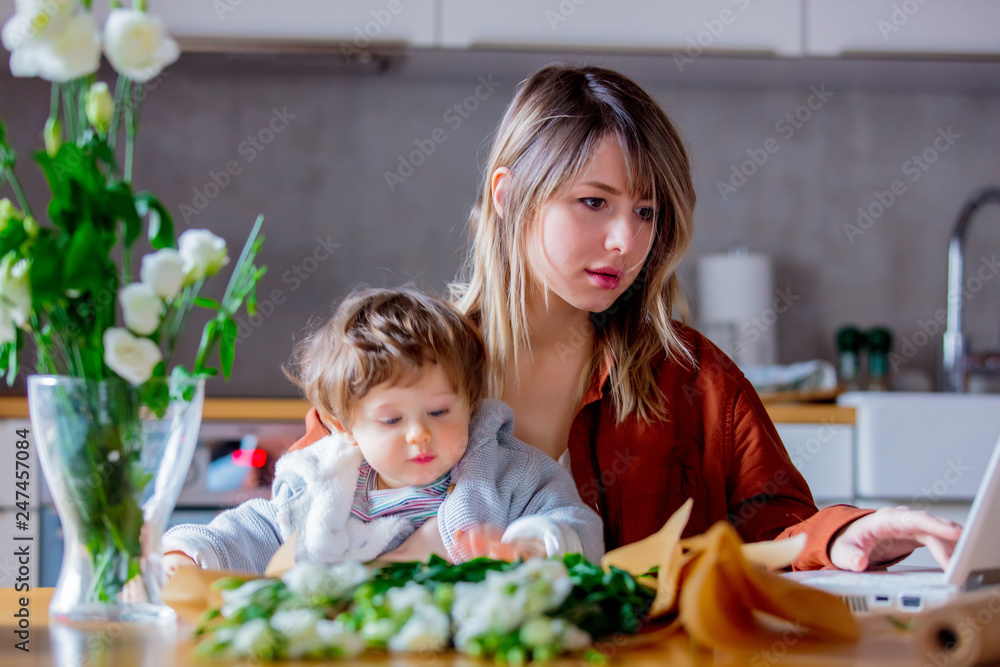 Mother with a child trying to working as a florist businesswoman and wrapping rose bouquet for sale via internet store. Located at home in a kitchen Mother with a child trying to working as a florist businesswoman and wrapping rose bouquet for sale via internet store. Located at home in a kitchen