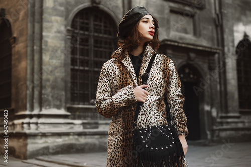 Outdoor fashion portrait of young woman wearing trendy leopard print coat, leather beret, holding black suede bag with fringe, posing in street of European city. Copy, empty space for text 