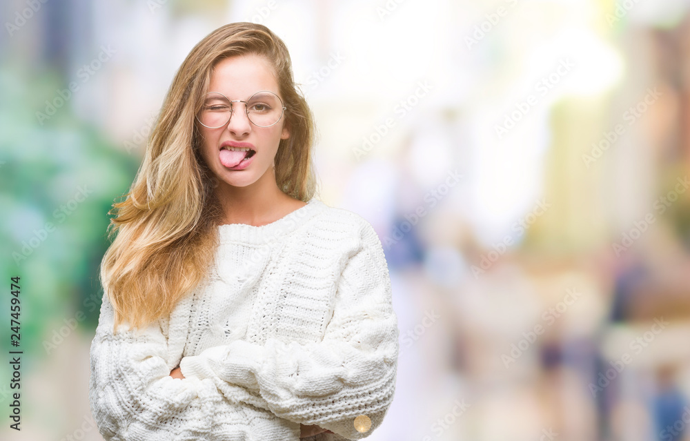 Young beautiful blonde woman wearing winter sweater and sunglasses over isolated background sticking tongue out happy with funny expression. Emotion concept.