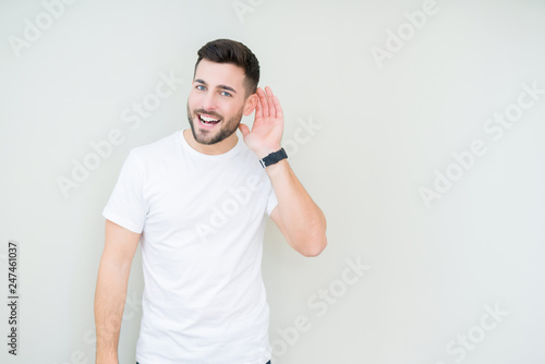 Young handsome man wearing casual white t-shirt over isolated background smiling with hand over ear listening an hearing to rumor or gossip. Deafness concept.