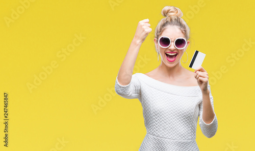 Young beautiful blonde woman holding credit card over isolated background annoyed and frustrated shouting with anger, crazy and yelling with raised hand, anger concept