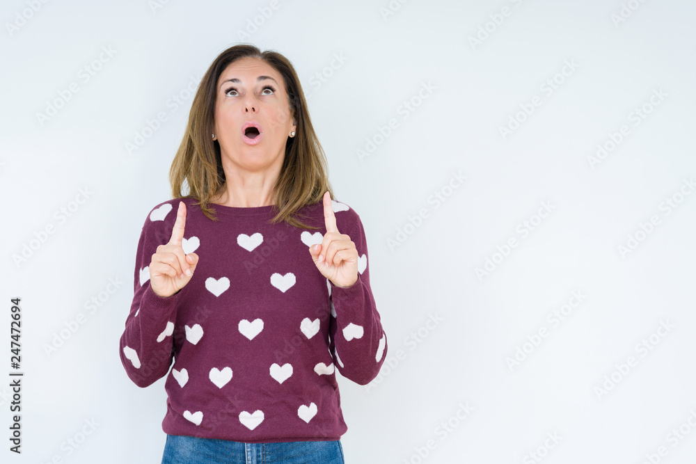 Beautiful middle age woman wearing heart sweater over isolated background amazed and surprised looking up and pointing with fingers and raised arms. Beautiful middle age woman wearing heart sweater over isolated background amazed and surprised looking up and pointing with fingers and raised arms.