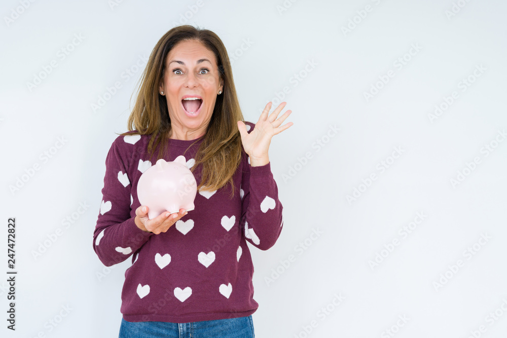 Middle age woman holding piggy bank over isolated background very happy and excited, winner expression celebrating victory screaming with big smile and raised hands Middle age woman holding piggy bank over isolated background very happy and excited, winner expression celebrating victory screaming with big smile and raised hands