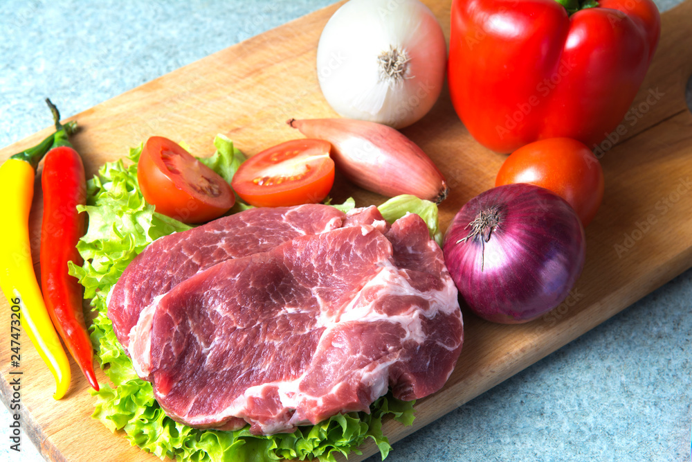Pork-neck meat steaks on lettuce on background of radishes, tomato, red chili peppers, yellow chili peppers, green paprika, yellow paprika, red paprika, black pepper. Horizontal. Red onions, shallots Pork-neck meat steaks on lettuce on background of radishes, tomato, red chili peppers, yellow chili peppers, green paprika, yellow paprika, red paprika, black pepper. Horizontal. Red onions, shallots