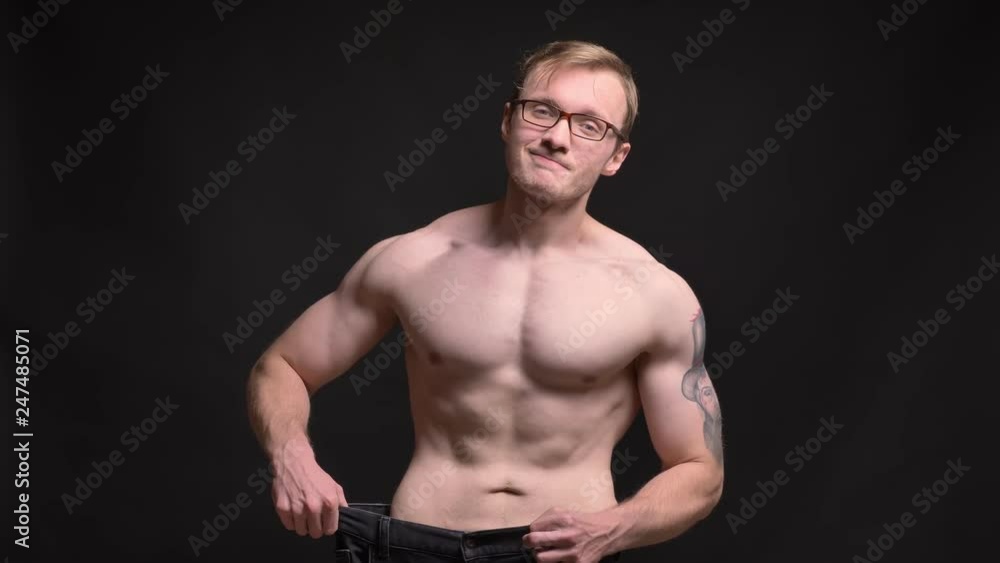 Portrait of naked young muscular man in glasses happily showing the results of losing weight into camera on black background. Portrait of naked young muscular man in glasses happily showing the results of losing weight into camera on black background.
