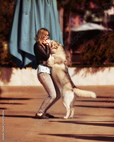 A grey Siberian husky female dog is staying on hind legs close to a young caucasian blond haired girl. A woman wears grey jeans, black jacket and light blue blouse. The background is brown.