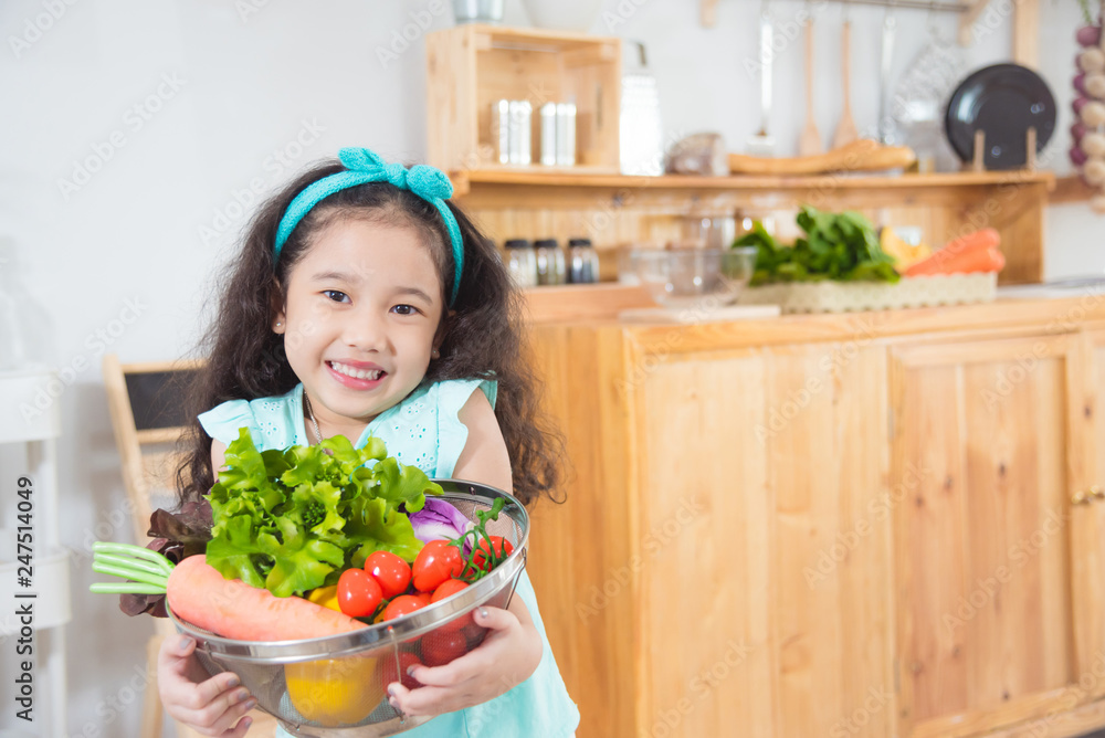 Little asian girl holding basket with many vegetables and smiles in kitchen Little asian girl holding basket with many vegetables and smiles in kitchen
