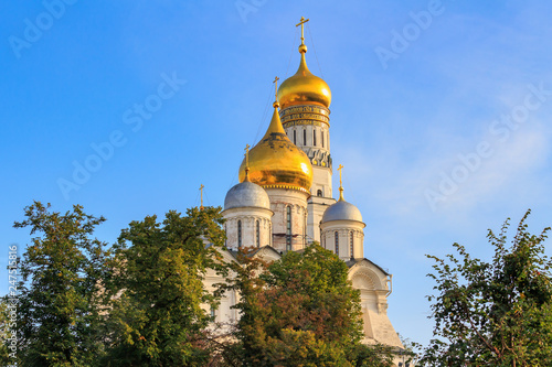 Cathedral of the Archangel and Ivan the Great Bell-Tower on Moscow Kremlin territory on a blue sky and green trees background in sunny morning