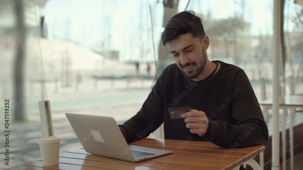 Happy young man holding credit card and using laptop. Handsome bearded young man shopping online via laptop computer and sitting with hands behind head and toothy smile in cafe. E-commerce concept Happy young man holding credit card and using laptop. Handsome bearded young man shopping online via laptop computer and sitting with hands behind head and toothy smile in cafe. E-commerce concept