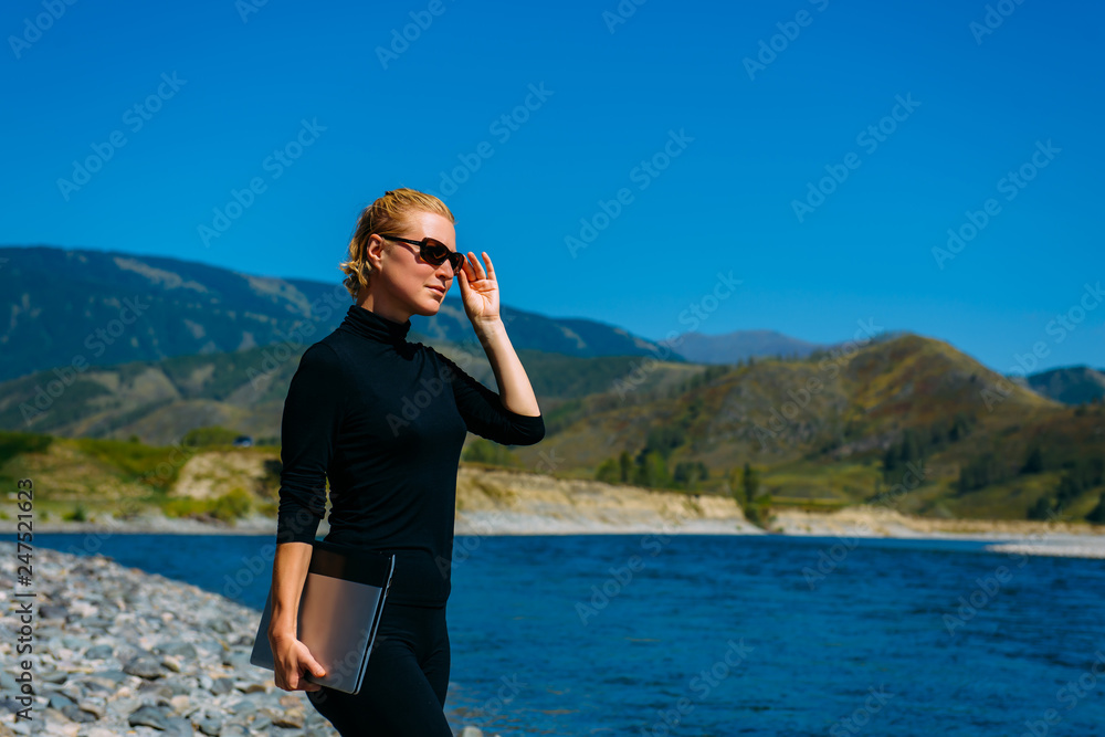 Freelancer with laptop on the nature. Blogger hipster traveler business lady pretty girl finished work outdoor. Young blondy woman with sunglasses in black standing next to mountain river Freelancer with laptop on the nature. Blogger hipster traveler business lady pretty girl finished work outdoor. Young blondy woman with sunglasses in black standing next to mountain river