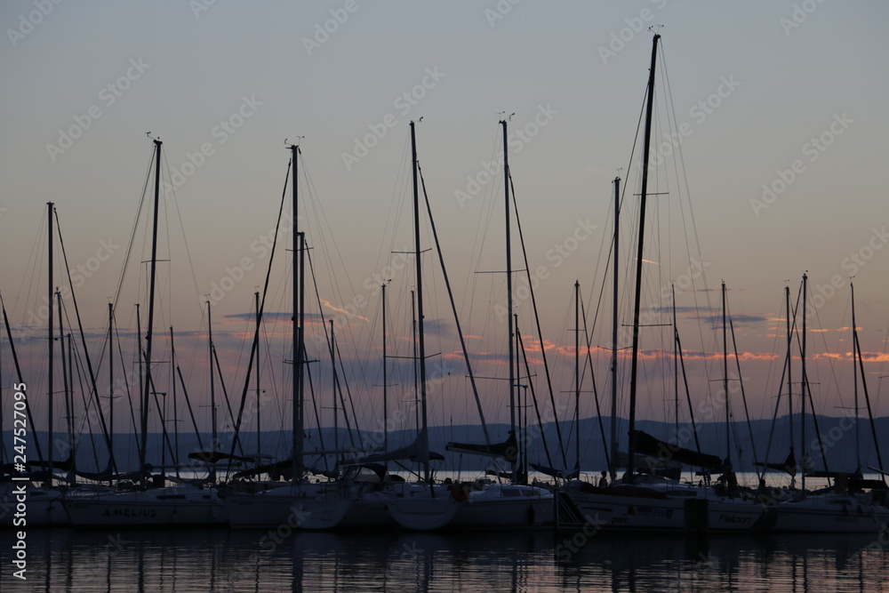A warm, pleasant summer evening at Lake Balaton. Yachts, sailboats, boats with masts in the marina, port. Mountains visible in the background. A beautiful twilight. Orange sky. A quiet end of the day.