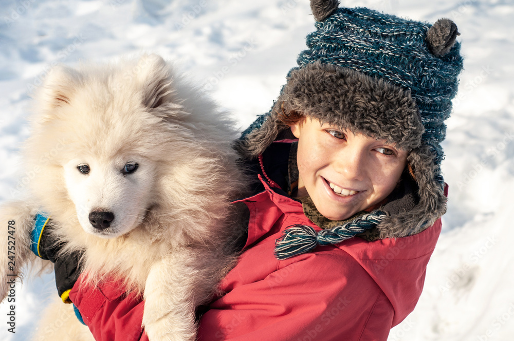 A boy in a hat with ears is hugging a samoyed puppy outdoors in
