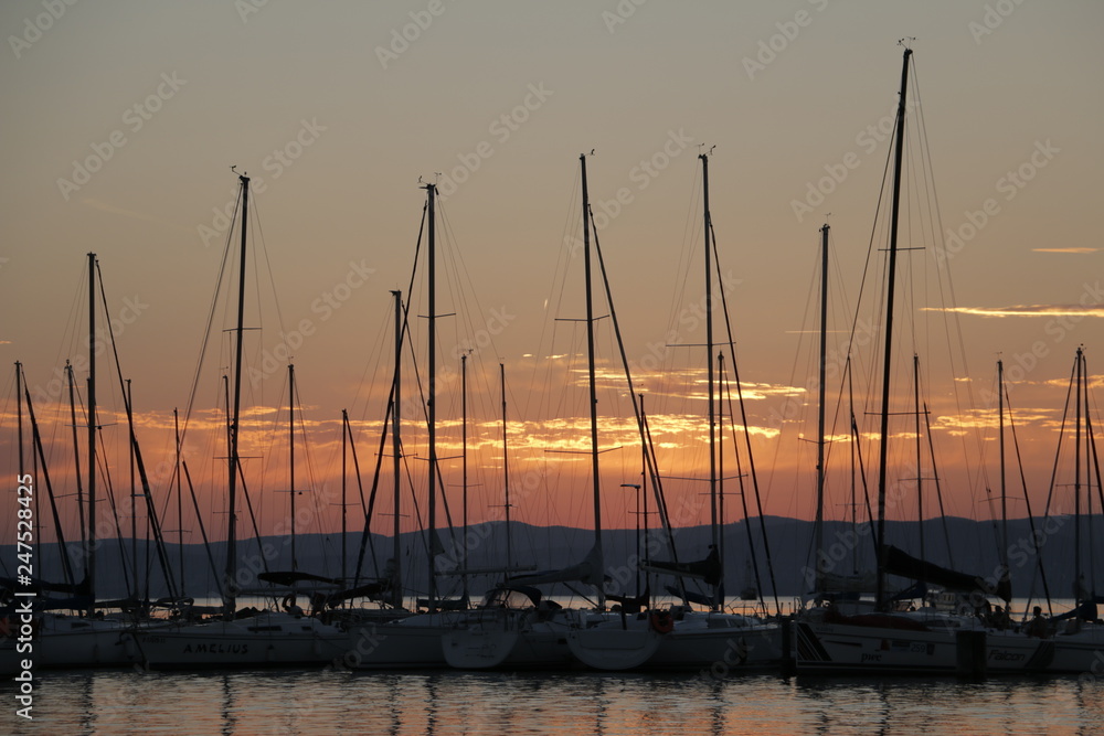 A warm, pleasant summer evening at Lake Balaton. Yachts, sailboats, boats with masts in the marina, port. Mountains visible in the background. A beautiful twilight. Orange sky. A quiet end of the day.
