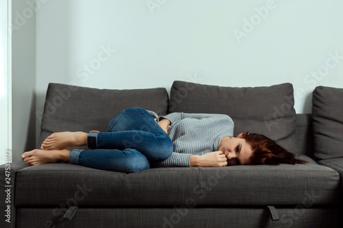 Young, beautiful girl suffers from stomach cramps and abdominal pain, lying on a home sofa. The concept of abdominal pain, female menstruation, stomach cramps.