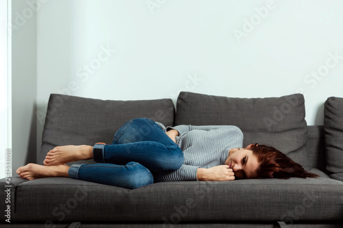 Young, beautiful girl suffers from stomach cramps and abdominal pain, lying on a home sofa. The concept of abdominal pain, female menstruation, stomach cramps.