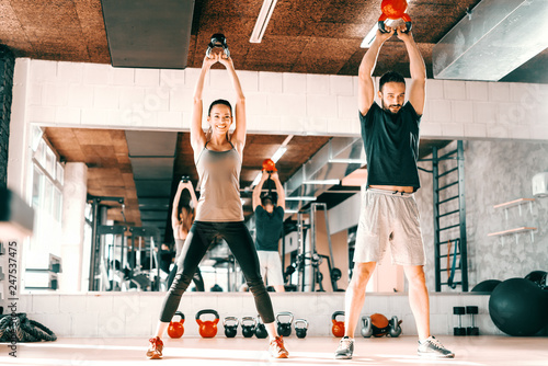 Happy Caucasian couple doing strength exercises with kettlebell while standing in gym. In background mirror with their reflection.