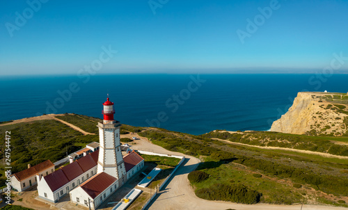 Aerial; drone view of white-red lighthouse Cabo Espichel on the edge of the earth; turquoise water of Atlantic ocean stretching to horizon; beautiful portuguese viewpoint with old historic sightseeing