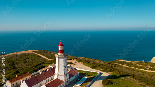 Aerial; drone view of white-red lighthouse Cabo Espichel on the edge of the earth; turquoise water of Atlantic ocean stretching to horizon; beautiful portuguese viewpoint with old historic sightseeing