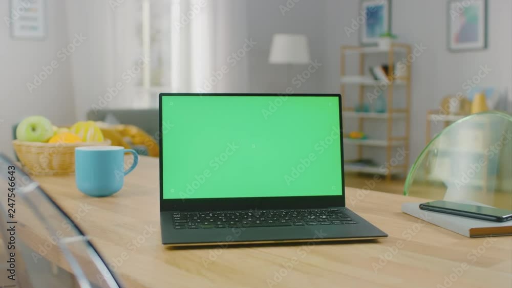 Shot of a Modern Laptop with a Horizontal Green Screen Mock Up on a Wooden Table at Home. Smartphone Lies on a Table Next to the Computer. Shot of a Modern Laptop with a Horizontal Green Screen Mock Up on a Wooden Table at Home. Smartphone Lies on a Table Next to the Computer.