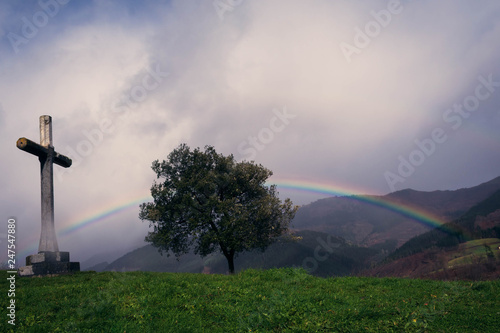 El arco iris sobre un árbol y saliendo desde la cruz. El día es nublado con tormentas.