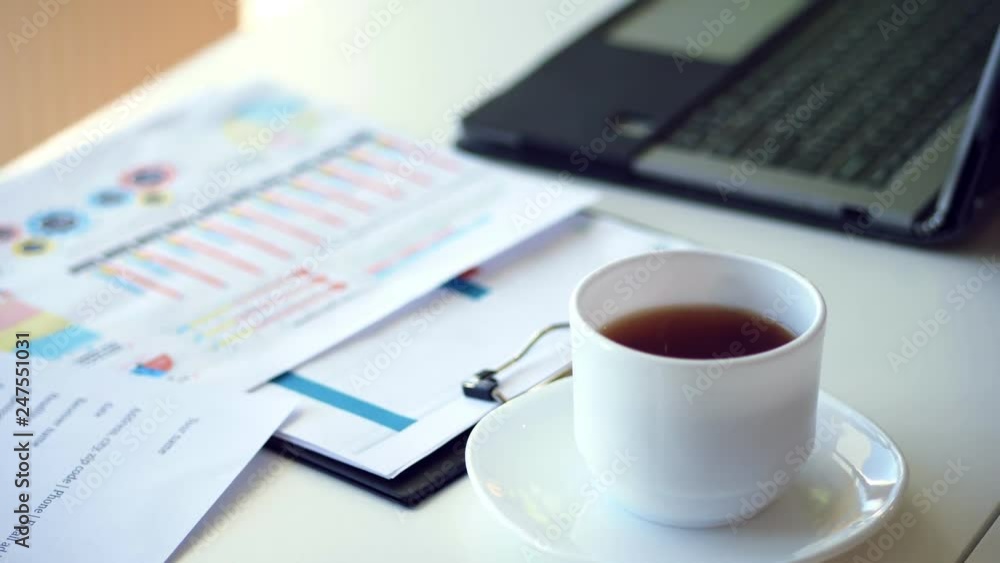 close up, on a white desktop there is a white cup of tea, next to a laptop and spread out business graphs close up, on a white desktop there is a white cup of tea, next to a laptop and spread out business graphs