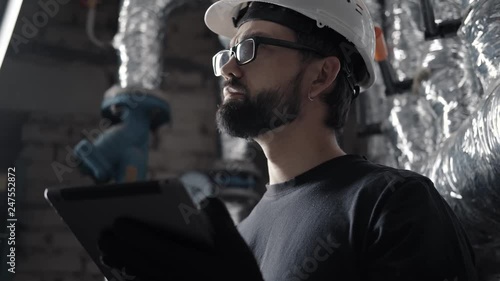 Wallpaper Mural Technician (engineer) in a helmet works in a boiler room with a tablet. On the background of hot pipes in the corrugation. A plumber does a ventilation or heating system check using a gadget. Torontodigital.ca