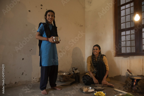 Happy rural woman sitting in kitchen cooking food on firewood on the floor while her school going daughter stands with a tiffin box.	 photo