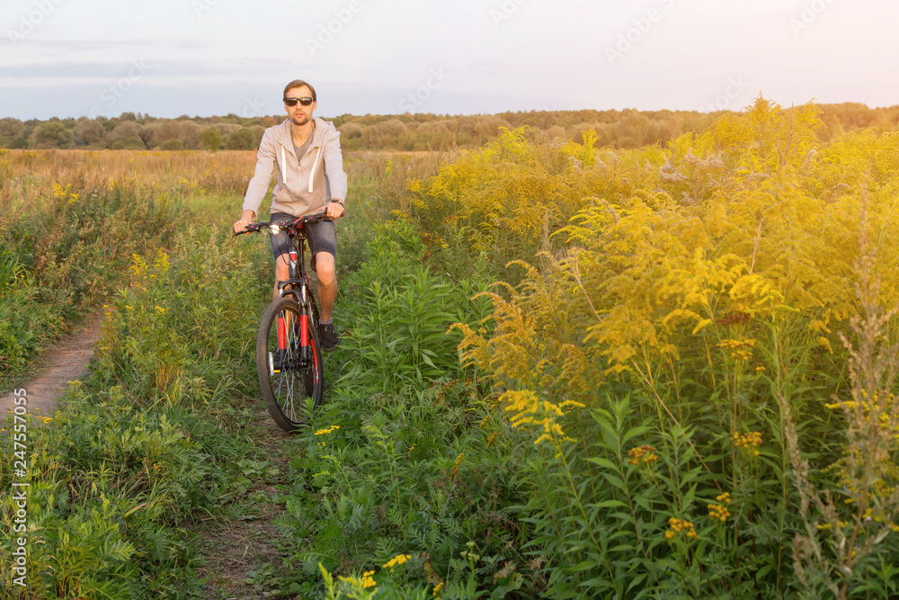 Fototapeta premium Young men on bike, cyclist in field with yellow flowers in sunlight, nature with copy space 