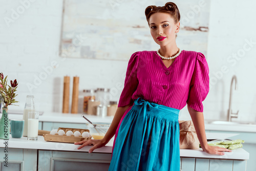 Stylish pin up girl in crimson dress and blue apron standing near kitchen table with various products