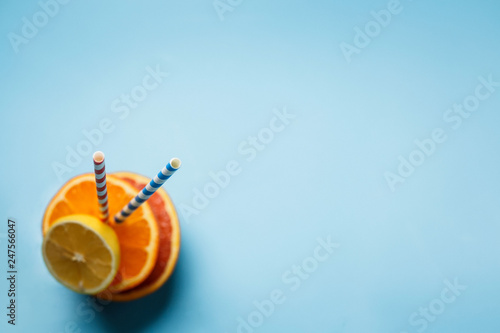 a glass of fresh juce and two paper tubes next to a lemon and an orange on a blue background with copy space