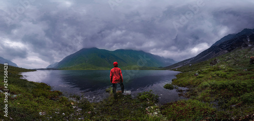 man stands by lake in red jacket in cloudy weather panorama