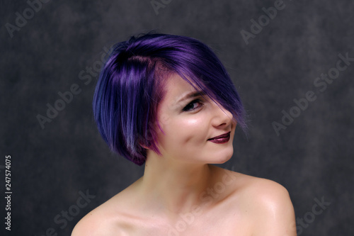 A beautiful, sexy girl with purple hair and a short haircut sits in the middle of the photo with a gray background