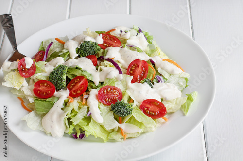 Tela Plate of homemade fresh salad with buttermilk ranch dressing, tomatoes, broccoli, cabbage and carrots served over a white wooden table