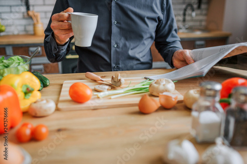 Cut view of man sitting at table in kitchen. He drink from white cup and read journal. Spices and colorful vegetables on table.