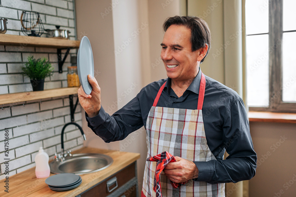 Cheerful positive man stand alone in kitchen. He old plate and look at it. Man smile. He dried it with towel.