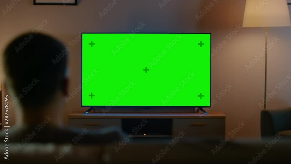 Young Man in Glasses is Sitting on a Sofa and Watching TV with Horizontal Green Screen Mock Up. It's Evening and Room at Home Has Working Lamps. Young Man in Glasses is Sitting on a Sofa and Watching TV with Horizontal Green Screen Mock Up. It's Evening and Room at Home Has Working Lamps.