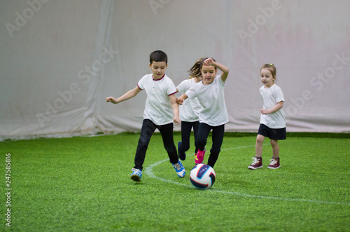 Children in white t-shirts playing football indoors. Kids running on the field after the ball