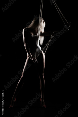 Beautiful slender blonde girl, wearing a white bodisuit and red stilettos, sensually plays and poses with the ropes. Black background. Artistic noir silhouette photo