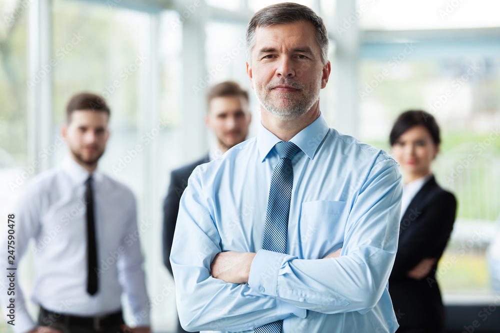 Modern mature businessman smiling and looking at camera with his colleagues in the background at office. Modern mature businessman smiling and looking at camera with his colleagues in the background at office.