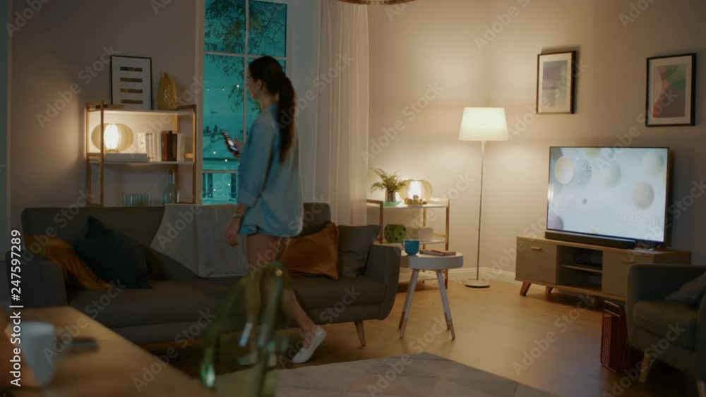 Young Beautiful Woman Gives a Voice Command to a Smart Home Application on Her Smartphone and Lights in the Room are Being Turned On. She Walks and Sits on a Couch. It's a Cozy Evening.