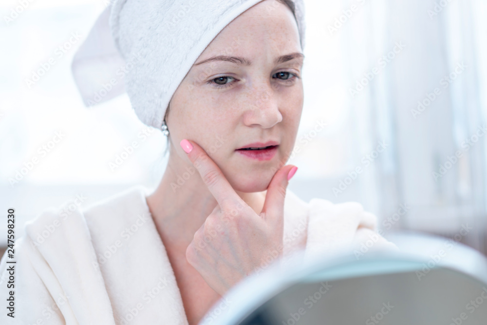 Beautiful happy young woman with a towel on her head looking at her skin in a mirror. Hygiene and care for the skin Beautiful happy young woman with a towel on her head looking at her skin in a mirror. Hygiene and care for the skin