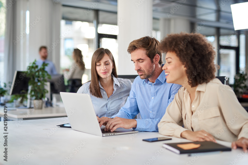 Coworkers sitting at table and solving problem. Man using laptop while two female colleagues looking at laptop and helping him..