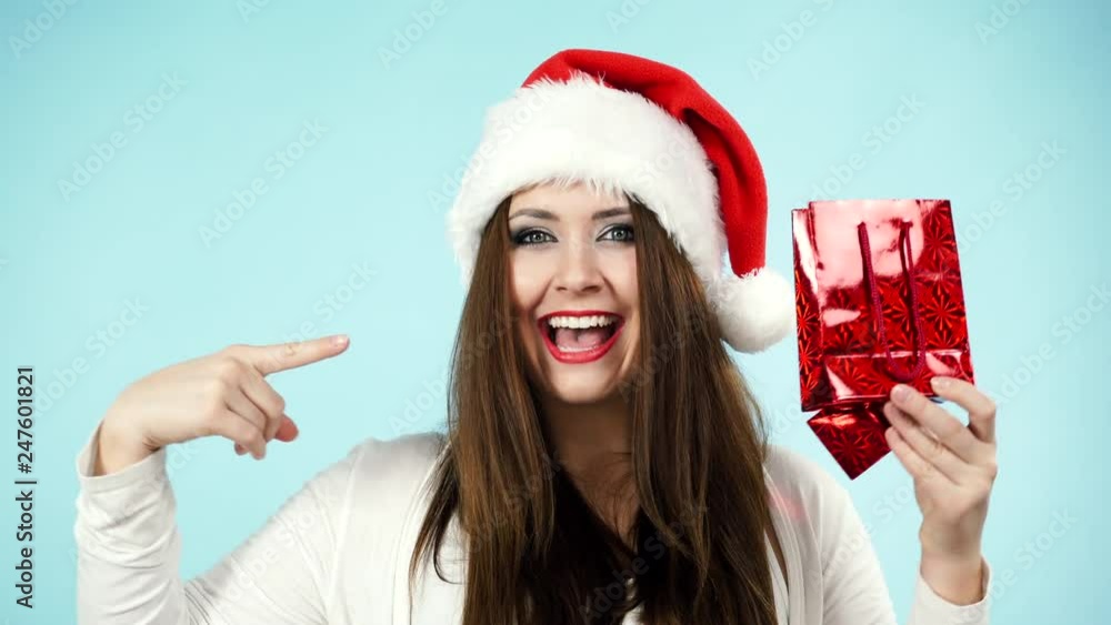 Happy smiling young woman in santa claus helper hat holding small red shopping bag, gift packaging, on blue. Christmas holidays Happy smiling young woman in santa claus helper hat holding small red shopping bag, gift packaging, on blue. Christmas holidays
