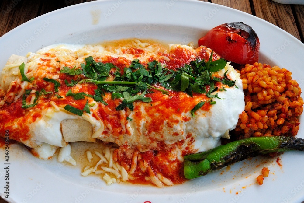 A plate of Turkish Beyti kebab consisting of ground beef, grilled on a skewer and served wrapped in lavash, topped with tomato sauce, yogurt and cheese with bulgur pilaf